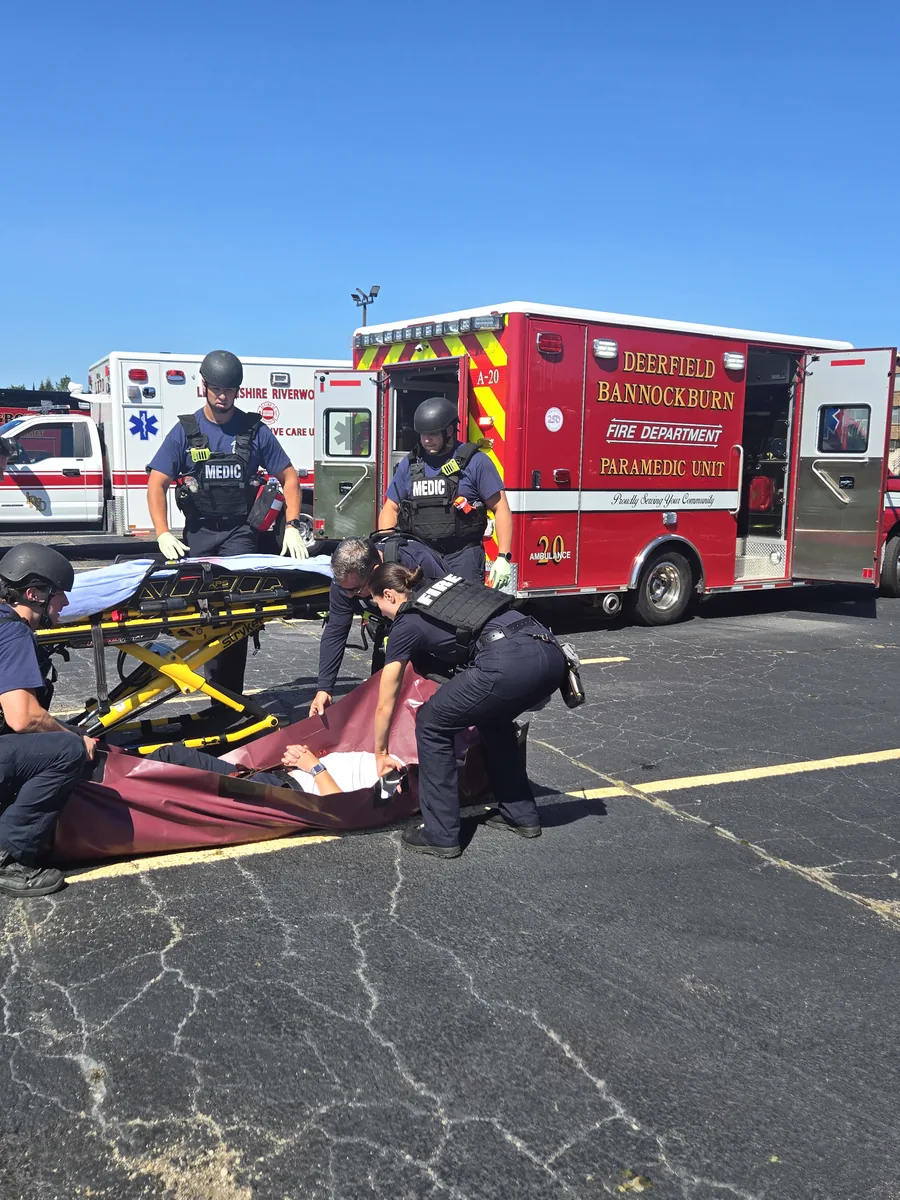 Highland Park EMS System providers and firefighters in tactical protective gear package a simulated patient during a drill, beside a Deerfield-Bannockburn Fire Department paramedic ambulance.