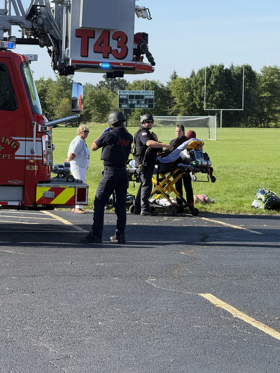 Responders in tactical protective gear tend to a simulated patient on a stretcher beside a Wheeling Fire Department aerial truck during a multi-agency drill at a sports field.